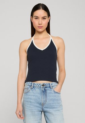 Jeune femme portant un haut noir à col licou avec une bordure blanche et un jean bleu clair taille haute, debout avec une main dans la poche.
