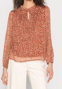 Blouse couleur rouille avec un motif floral, décolleté en forme de trou de serrure, manches longues et un tissu léger et transparent.