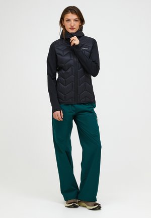 Kvinde iført en sort quiltet jakke, grønblå cargobukser og brune sneakers, der står med den ene hånd ved hagen mod en ensfarvet baggrund.