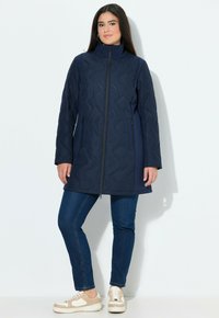 Kvinna står mot en vit bakgrund iklädd en marinblå vadderad jacka, blå jeans och beigea samt vita sneakers.