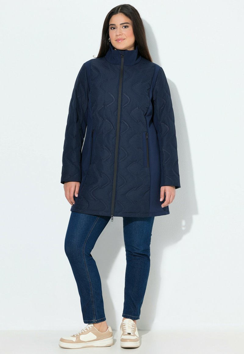 Kvinna står mot en vit bakgrund iklädd en marinblå vadderad jacka, blå jeans och beigea samt vita sneakers.