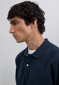 Młody mężczyzna z kręconymi ciemnymi włosami, ubrany w granatową koszulę z kołnierzykiem i białą koszulkę pod spodem, zwrócony w lewo na jednolitym tle.