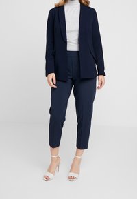 Veste de blazer bleu marine à col châle, associée à un pantalon court assorti et des talons ouverts couleur nude. Tissu lisse avec une coupe ajustée.