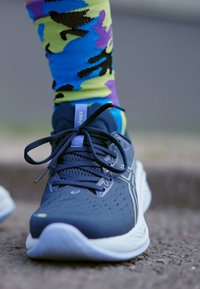 Sneaker con una toma in rete blu navy scuro, suola grigia chiara e dettagli riflettenti. Abbinato a calzini con un motivo camouflage colorato in blu, verde e viola.