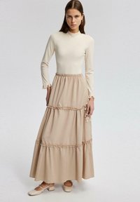 Beige langemouwen top met een gestructureerd lijf en gerimpelde manchetten, gecombineerd met een meerlaagse beige rok met geplooide details en enkelschoenen.