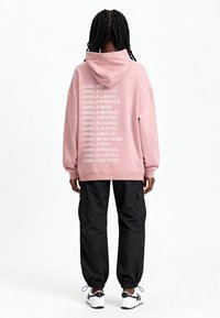 Rózsaszín kapucnis pulóver, fehér felirattal a hátsó oldalán, fekete cargo nadrág, és fehér sportcipő. A pulóver laza fazonú, állítható húzózsinórral.