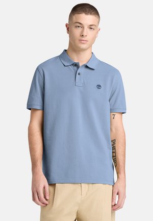 Jonge man draagt een lichtblauw polohemd met korte mouwen en beige broek, staand tegen een effen lichte achtergrond.