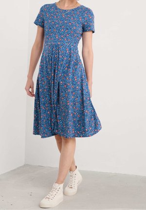 Vrouw draagt een blauwe bloemenjurk tot op de knie met korte mouwen en witte hoge sneakers, staand tegen een witte muur.