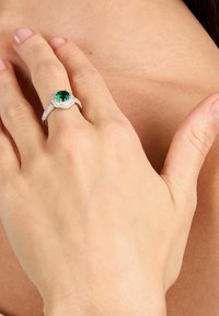 Anello in smeraldo con una pietra verde tonda, circondata da piccoli diamanti, montata su un band in argento lucido con un design intrecciato.
