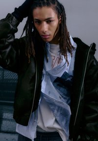 Svart bomberjacka i läder med ribbade muddar, lager-på-lager över en blårandig skjorta och en vit T-shirt, med text- och grafiska accenter.