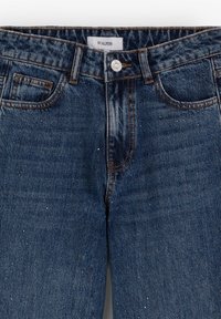 Jean en denim bleu foncé avec poches avant, passants de ceinture, bouton en métal et étiquette de marque visible "Scalpers" à l'intérieur de la ceinture.