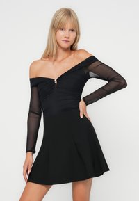 Vestuário preto com um design à mostra de ombros, mangas longas transparentes, um corpete justo com franzidos e uma saia rodada. Acento em metal na gola.