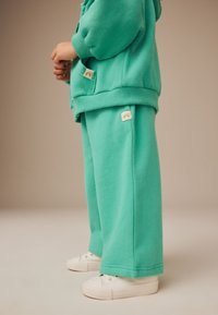 Turquoise sweatshirt and matching sweatpants made of soft fabric, featuring pockets and small rainbow logo patches, paired with white sneakers.