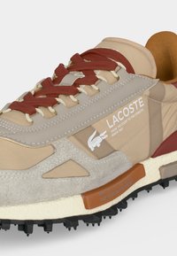 Μπεζ και ανοιχτό καφέ αθλητικό παπούτσι Lacoste με κόκκινα κορδόνια, υφή σόλας και λευκό λογότυπο που γράφει "From the court to the streets since 1933."