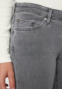 Harmaat farkut, joissa on klassinen viiden taskun muotoilu, hienovarainen tekstuuri ja metalliset yksityiskohdat, sekä ompelut yksityiskohdat saumoissa.