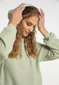 Felpa verde chiaro a trama waffle con cappuccio regolabile, taglio morbido e maniche lunghe. Il materiale sembra morbido e accogliente.