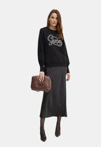 Pullover nero con logo "Guess" in argento, abbinato a una lunga gonna nera di raso, mentre tiene una borsa in pelle marrone con tracolla a catena.