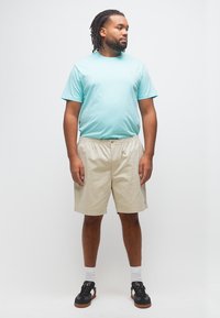 Άνδρας με πλεξούδες, φορώντας ανοιχτό γαλάζιο μπλουζάκι, μπεζ σορτς, άσπρες κάλτσες και μαύρα αθλητικά παπούτσια, στέκεται μπροστά σε απλό λευκό φόντο.