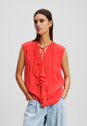 Red sleeveless blouse with ruffled front detail, keyhole neckline, and soft fabric. Paired with light blue high-waisted jeans.