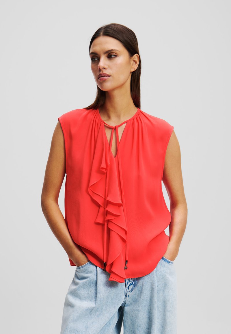 Blusa sin mangas roja con detalle de volantes en la parte delantera, escote en forma de ojo de cerradura y tela suave. Combinada con unos jeans de cintura alta en azul claro.