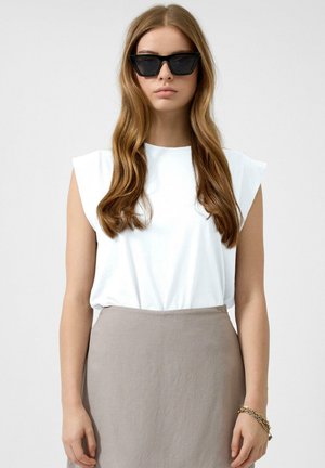 Vrouw met lang golvend haar, draagt zwarte cat-eye zonnebril, een witte mouwloze top, een grijze rok en een gouden kettingarmband.