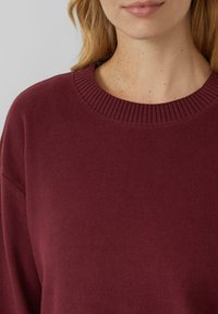 Vrouw in een bordeauxrode gebreide trui met geribbelde ronde halslijn, die haar nek en onderkant van het gezicht toont met een lichte huid en sproeten.