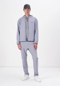 Grå treningsjakke med hette og glidelås, kombinert med matchende grå bukser. Funksjoner inkluderer et minimalistisk design og hvite sneakers.