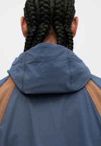 Gewatteerde jas van blauwe stof met bruine accenten op de schouders. Geweven materiaal met zichtbare stiksels bij de hals en naden.