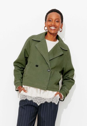 Femme souriante portant une veste verte courte, un haut blanc orné de dentelle et un pantalon bleu marine à fines rayures, les mains dans les poches, sur fond blanc.