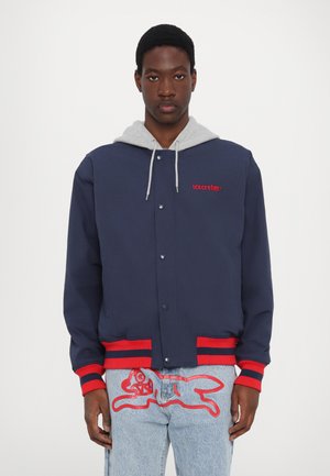 Navy blue jacket with gray hood, red accents, and snap buttons; worn with light blue jeans featuring red embroidered design.