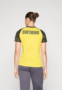 T-shirt de sport jaune avec des manches à motifs noirs. Le dos présente "DORTMUND" en lettres grasses noires. Texture lisse, design ajusté.
