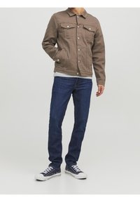 Hnědá džínová bunda se dvěma náprsními kapsami a knoflíkovým zapínáním, v kombinaci s tmavě modrými džíny a námořnickými teniskami. Pod bundou je pruhovaná košile.