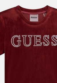 Burgundská sametová tričko s nápisem "GUESS" zdobeným průhlednými kamínky, kulatým výstřihem a krátkými rukávy. Měkká textura.