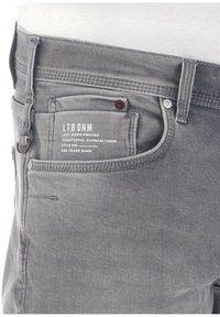 Los jeans de mezclilla gris cuentan con un bolsillo trasero etiquetado, cierre de botón y una textura distintiva, con sutiles detalles de costura que realzan el diseño.