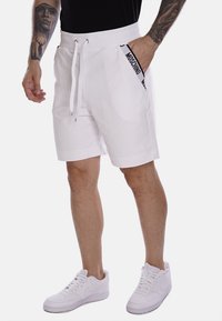 Pantaloni corti bianchi con vita regolabile, caratterizzati da un logo "Moschino" nero lungo il lato. Indossati con sneakers bianche, texture del tessuto liscia.