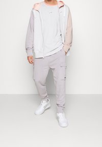 Chaqueta con capucha en gris y beige con mangas en contraste, combinada con pantalones cargo grises. Zapatillas blancas completan el conjunto.