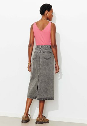 Femme portant un haut rose sans manches, une jupe midi en denim gris délavé taille haute avec une fente arrière, et des chaussures à lacets imprimé léopard, debout face au mur.