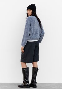 Pull en maille bleu, short gris à rayures, et bottes en cuir noires jusqu'au genou avec des boucles. Accompagné d'un bonnet noir.