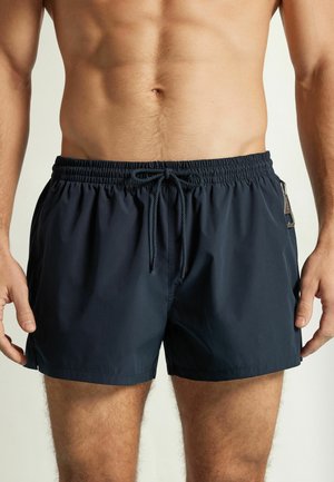 Torse masculin et parties supérieures des jambes portant un short de bain noir avec taille élastique et cordon, debout devant un fond neutre.