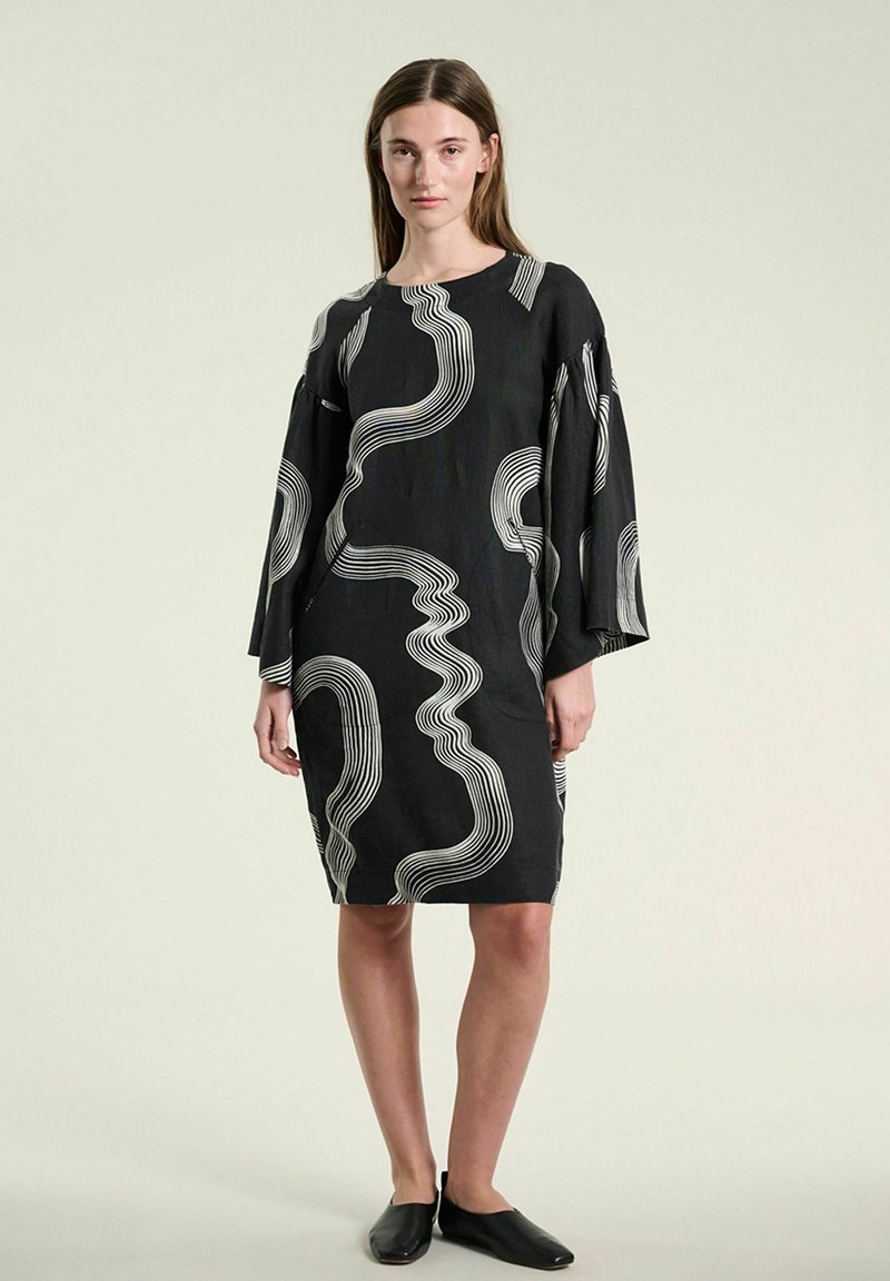 Femme portant une robe noire longueur genou avec des motifs abstraits de lignes ondulées blanches et des manches larges, associée à des chaussures plates noires.