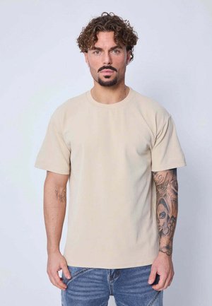 T-shirt beige à manches courtes avec un col rond et une texture lisse. Tatouages décoratifs visibles sur le bras gauche, porté avec un jean bleu.