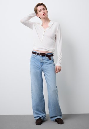 Camiseta blanca de manga larga con cuello, combinada con vaqueros anchos de pierna azul claro y cinturón marrón. La modelo lleva zapatos marrón oscuro.