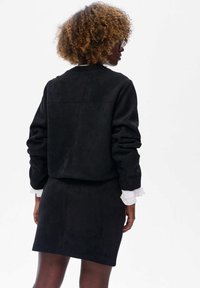 Kobieta z kręconymi włosami, ubrana w czarną kurtkę i spódnicę, z mankietami białej koszuli, zwrócona tyłem na białym tle.