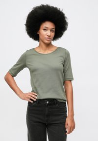 Γυναίκα με αφρο μαλλιά, φορώντας ελαιόχρωμο κοντομάνικο πουκάμισο και μαύρα τζιν, στέκεται με το ένα χέρι στη μέση της σε απλό φόντο.