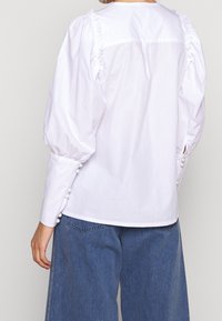 Blusa bianca a maniche a sbuffo con dettagli arricciati sul retro. Presenta accenti in perla sui polsini. Abbinata a pantaloni in denim blu a gamba larga.