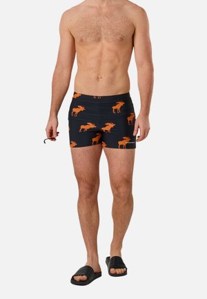 Mand iført sorte badetrusser med orange elgmønster, sorte slidesandaler, holder sort snor, stående mod en hvid baggrund.