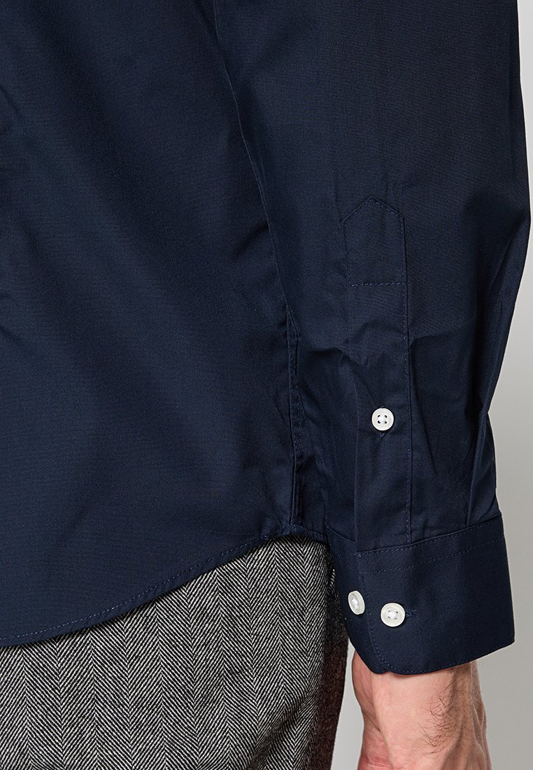 Polsino di camicia a maniche lunghe blu navy con due bottoni bianchi indossato con pantaloni grigi a motivo spina di pesce, che mostrano i dettagli della cucitura laterale.