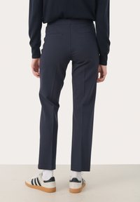 Pantalons sur mesure bleu marine avec une jambe droite, deux poches arrière et une texture lisse, associés à des baskets blanches et des chaussettes côtelées.
