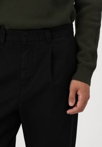 Svarta veckade byxor i vävt tyg, med en medelhög midja, sidofickor och en slät yta. Hand som vilar på midjebandet.