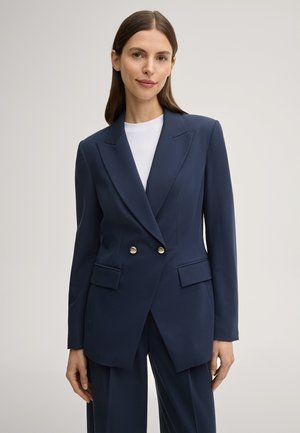 Vrouw met stijlbruin haar draagt een marineblauw dubbelreeksblazer over een wit shirt met ronde hals, staand tegen een effen lichte achtergrond.